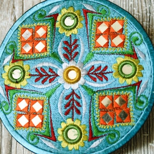 Op de afbeelding: Een ronde, blauwe, geborduurde wandhanger met een bloemenmotief en gespiegelde accenten. Het ontwerp heeft een centrale bloem met vier kleinere bloemen eromheen. De bloemen zijn geborduurd in tinten geel, groen, rood en wit. De gespiegelde accenten zijn vierkant en symmetrisch gerangschikt.