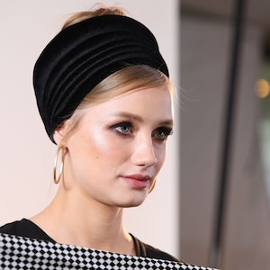 Op de afbeelding: Een vrouw met een zwart fluwelen hoofdband en grote gouden oorringen houdt een zwart-wit geblokt doek vast.