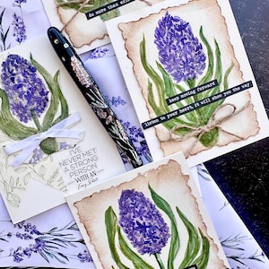 May include: A collection of handmade greeting cards featuring watercolor paintings of purple hyacinth flowers with green leaves. Each card has a unique quote, and the cards are tied with twine. A decorative pen with floral accents is also present.