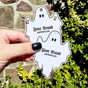 May include: Two white ghost-shaped earring display cards with the text "Your Brand" and "yourbrand.com". One card has a gold stud earring, the other has silver hoop earrings. The background is a stone wall and green foliage.