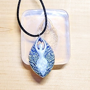 May include: A blue and white pendant with a goddess design, hanging on a black cord. The pendant is in front of a clear mold with the text "WHIMSICAL GODDESS MOLD". The pendant has a teardrop shape with intricate details.