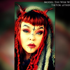 Puede incluir: Una mujer con cabello rojo y una peluca trenzada roja y blanca con cuernos, usando delineador de ojos negro y lápiz labial rojo, con marcas tribales negras y blancas en la cara.