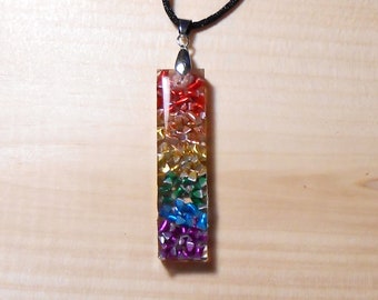 Puede incluir: Un collar con un colgante de arcoíris y una cadena de plata. El colgante está hecho de resina transparente llena de pequeñas piezas coloridas e iridiscentes.