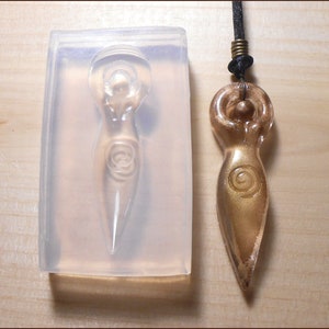 May include: A clear silicone mold for making a pendant in the shape of a female figure with a spiral design. The mold is next to a gold-colored pendant made from the mold.