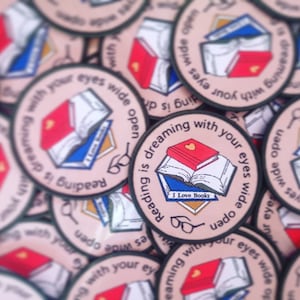 May include: A close-up of several round, pink and green embroidered patches with the text "Reading is dreaming with your eyes wide open" and a small red heart. The patches feature a stack of books with a red book on top and the text "I Love Books".