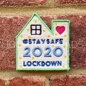 May include: A white house-shaped embroidered patch with a green border. The patch has a small window, a pink heart, and the text "Stay Safe 2020 Lockdown".