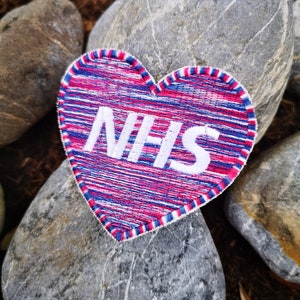 May include: A red, white and blue heart-shaped patch with the word "NHS" embroidered in white. The patch is sitting on a grey rock.