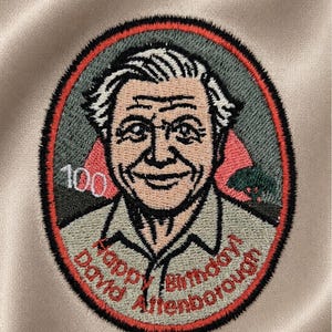 May include: Embroidered patch featuring a portrait of David Attenborough, with the text "Happy Birthday! David Attenborough" and the number "100". The patch has a red and black border and is set against a beige fabric.