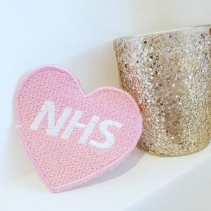 May include: Pink felt heart-shaped patch with white embroidered text that reads "NHS".