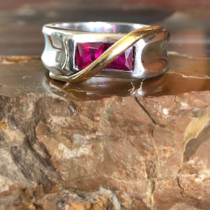 Op de afbeelding: Een zilveren en gouden ring met twee roze vierkante edelstenen. De ring ligt op een bruine steen.