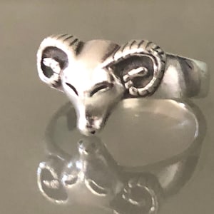 Może przedstawiać: Srebrny pierścionek z motywem głowy barana. Głowa barana jest szczegółowo wykonana z rogami i fakturowaną twarzą.