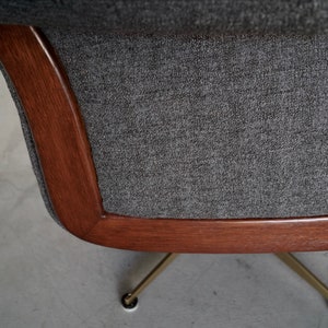 May include: Close-up of a mid-century modern chair. The chair features a dark gray fabric seat and back, framed by a curved, warm-toned wooden border. The chair rests on brass-colored legs.