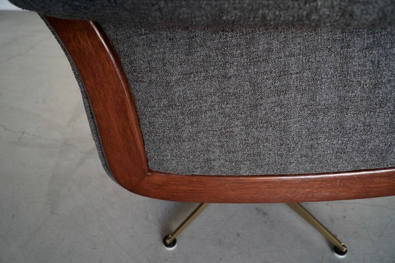 May include: Close-up of a mid-century modern chair. The chair features a dark gray fabric seat and back, framed by a curved, warm-toned wooden border. The chair rests on brass-colored legs.