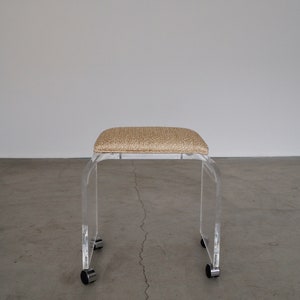 May include: A clear acrylic stool with a rectangular, beige woven seat. The stool has a curved, U-shaped design and sits on four small, rolling casters. The design is modern and minimalist.