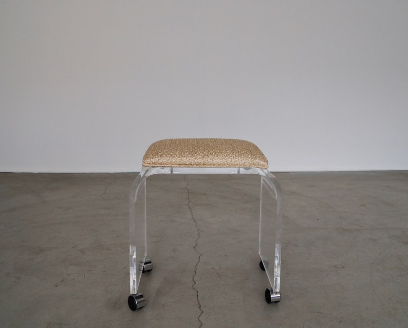 May include: A clear acrylic stool with a rectangular, beige woven seat. The stool has a curved, U-shaped design and sits on four small, rolling casters. The design is modern and minimalist.