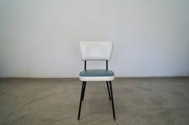 May include: A retro-style chair with a white backrest and a teal seat. The chair has a black metal frame and legs. The seat is trimmed with white. The chair is a vintage piece, likely from the mid-century modern era.
