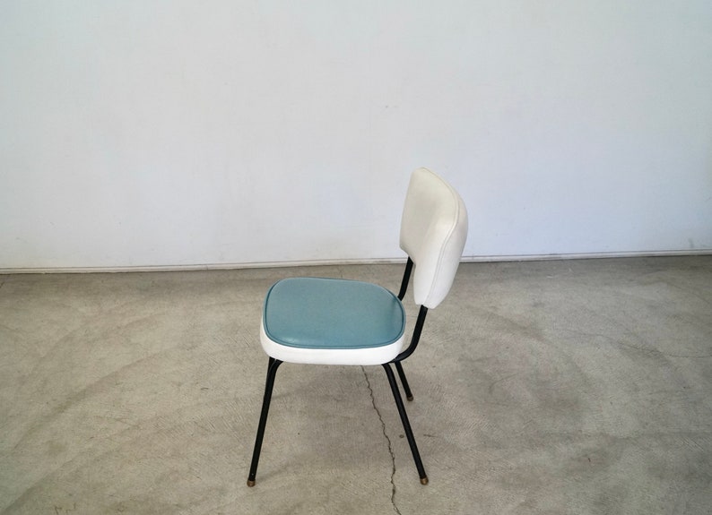 May include: A retro-style chair with a white back and a teal seat. The chair has a black metal frame and legs. The seat and back are upholstered. The chair is a vintage piece, perfect for a kitchen or dining room.