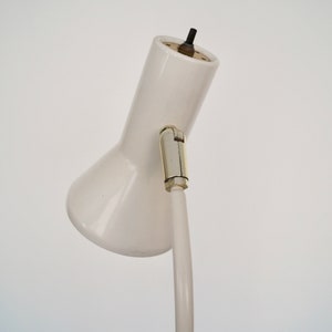 May include: A white desk lamp with a curved, adjustable arm and a conical shade. The lamp has a metal joint and a black power cord. The lamp is set against a plain white background.