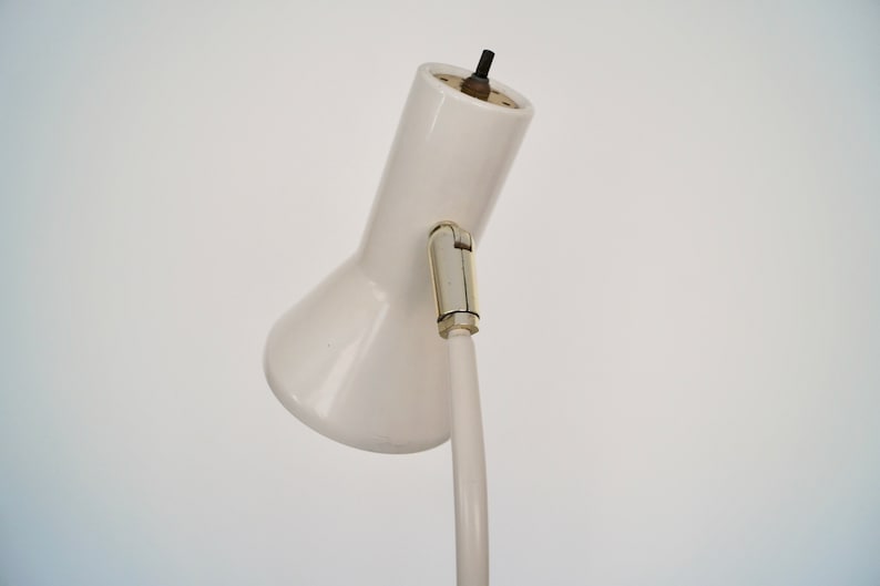 May include: A white desk lamp with a curved, adjustable arm and a conical shade. The lamp has a metal joint and a black power cord. The lamp is set against a plain white background.