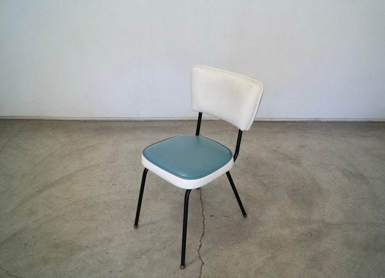 May include: A retro-style chair with a white backrest and a blue seat, outlined in white. The chair has black metal legs and a curved back support. The design is reminiscent of mid-century modern furniture.
