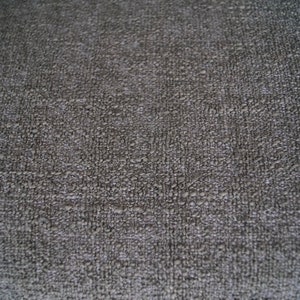 May include: Close-up of a textured, dark gray fabric. The surface has a slightly uneven, bumpy appearance, with a subtle pattern of small, raised elements. The overall tone is muted and neutral.