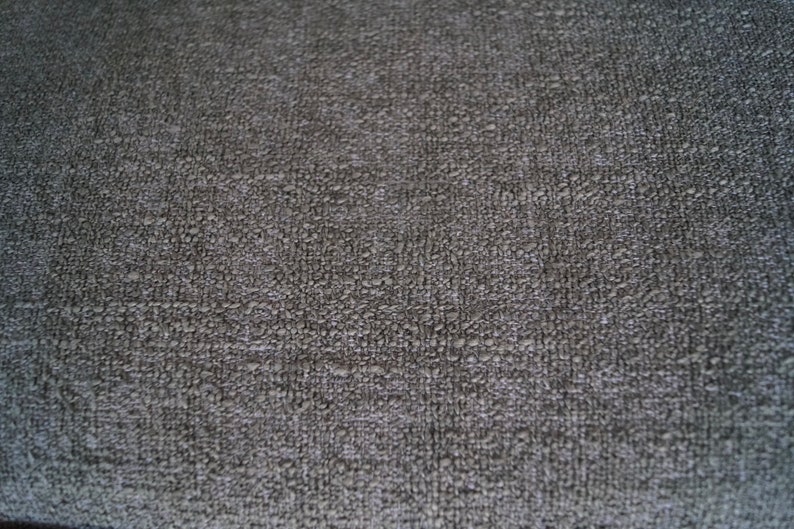 May include: Close-up of a textured, dark gray fabric. The surface has a slightly uneven, bumpy appearance, with a subtle pattern of small, raised elements. The overall tone is muted and neutral.
