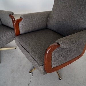 May include: Two gray upholstered armchairs with wooden armrests and brass-colored legs. The chairs have a mid-century modern design, with a curved back and armrests. The fabric is a textured gray, and the wood is a dark brown.