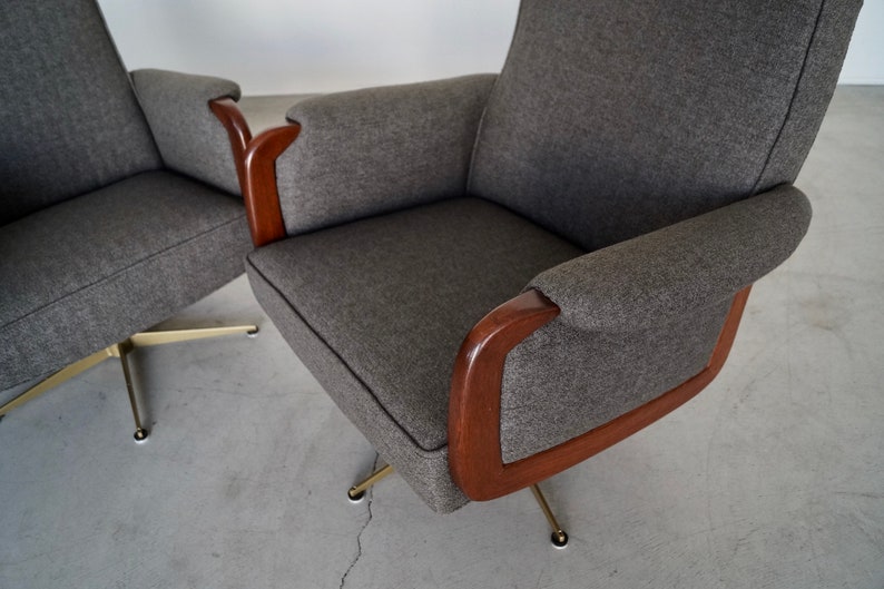 May include: Two gray upholstered armchairs with wooden armrests and brass-colored legs. The chairs have a mid-century modern design, with a curved back and armrests. The fabric is a textured gray, and the wood is a dark brown.