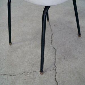 May include: A close-up of a chair with black metal legs and a white seat. The legs are thin and slightly curved, with brass-colored tips. The chair is set against a gray concrete surface.