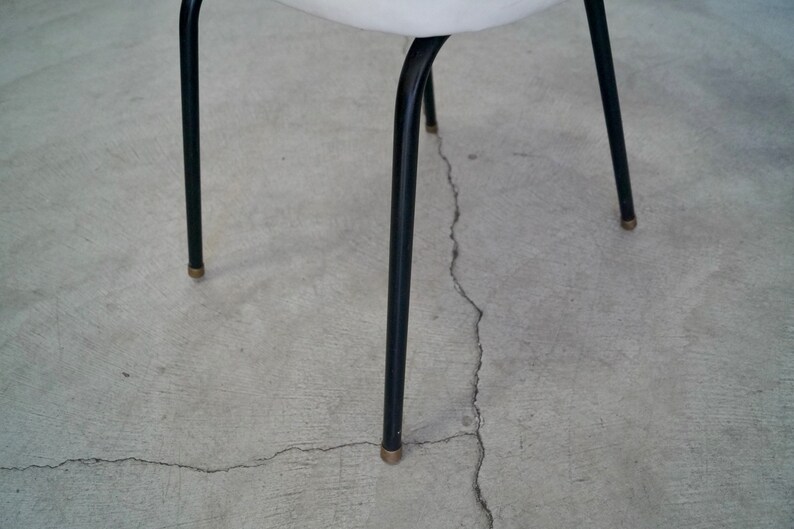 May include: A close-up of a chair with black metal legs and a white seat. The legs are thin and slightly curved, with brass-colored tips. The chair is set against a gray concrete surface.