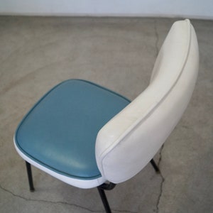 May include: A retro-style chair with a blue seat and white backrest. The seat is square with rounded corners, and the backrest curves upwards. The chair has black metal legs, and the design is reminiscent of mid-century modern furniture.
