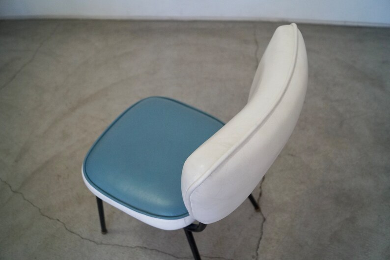 May include: A retro-style chair with a blue seat and white backrest. The seat is square with rounded corners, and the backrest curves upwards. The chair has black metal legs, and the design is reminiscent of mid-century modern furniture.