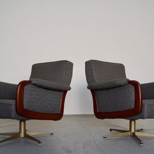 May include: Two mid-century modern swivel armchairs. The chairs have gray upholstery, dark wood armrests, and brass-colored star-shaped bases. The chairs are set against a plain white wall.