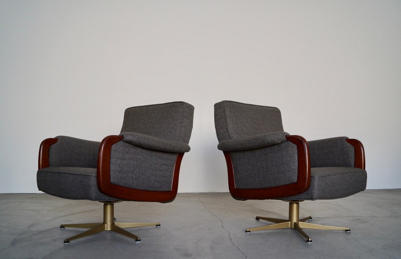 May include: Two mid-century modern swivel armchairs. The chairs have gray upholstery, dark wood armrests, and brass-colored star-shaped bases. The chairs are set against a plain white wall.