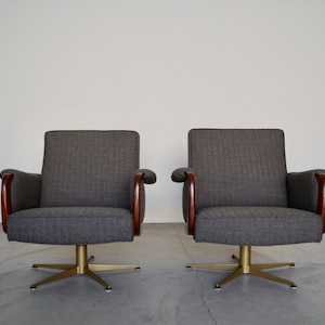May include: Two gray upholstered armchairs with wooden armrests and brass starburst bases. The chairs have a mid-century modern design, with a simple, clean aesthetic. The chairs are set against a plain white wall.