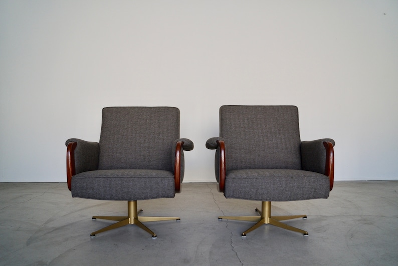May include: Two gray upholstered armchairs with wooden armrests and brass starburst bases. The chairs have a mid-century modern design, with a simple, clean aesthetic. The chairs are set against a plain white wall.