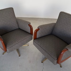 May include: Two gray upholstered armchairs with wooden armrests and brass-colored swivel bases. The chairs have a mid-century modern design, with a simple, clean aesthetic. The fabric appears to be a textured weave.