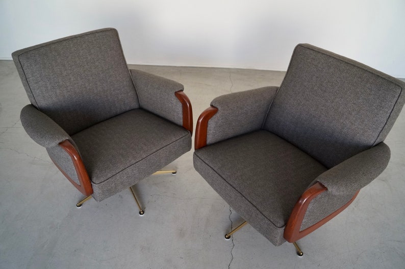 May include: Two gray upholstered armchairs with wooden armrests and brass-colored swivel bases. The chairs have a mid-century modern design, with a simple, clean aesthetic. The fabric appears to be a textured weave.