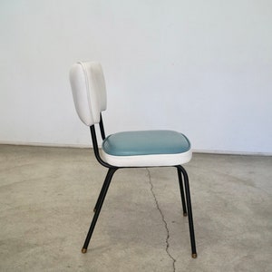 May include: A retro-style chair with a white backrest and a blue seat. The chair has a black metal frame and legs with brass-colored tips. The chair is a vintage piece.
