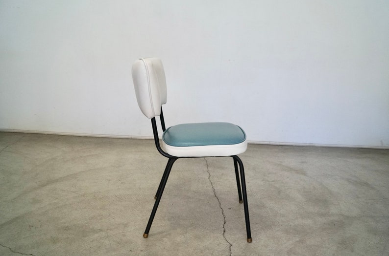 May include: A retro-style chair with a white backrest and a blue seat. The chair has a black metal frame and legs with brass-colored tips. The chair is a vintage piece.