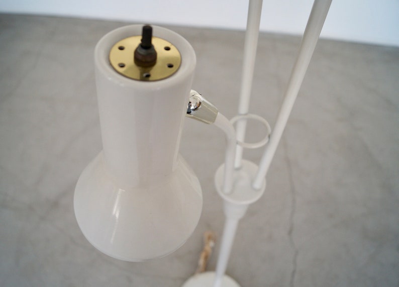 May include: A white, mid-century modern style floor lamp. The lamp features a rounded shade, a brass-colored top, and a slender, curved arm. The lamp's base and supporting structure are white.