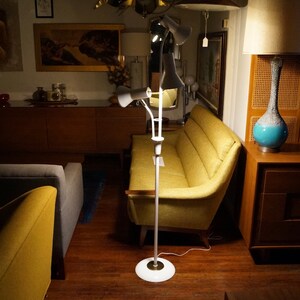 May include: A white and brass mid-century modern floor lamp with three adjustable cone-shaped shades. The lamp is standing on a white circular base. The background includes a yellow sofa and wooden furniture.
