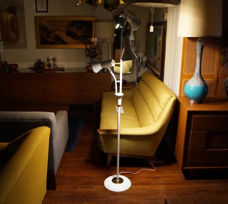 May include: A white and brass mid-century modern floor lamp with three adjustable cone-shaped shades. The lamp is standing on a white circular base. The background includes a yellow sofa and wooden furniture.