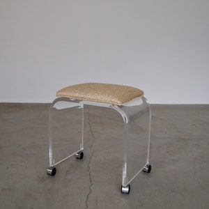 May include: Clear acrylic stool with a rectangular, cushioned seat in a speckled tan fabric. The stool has a curved design and sits on four small wheels for easy movement. A modern piece of furniture.
