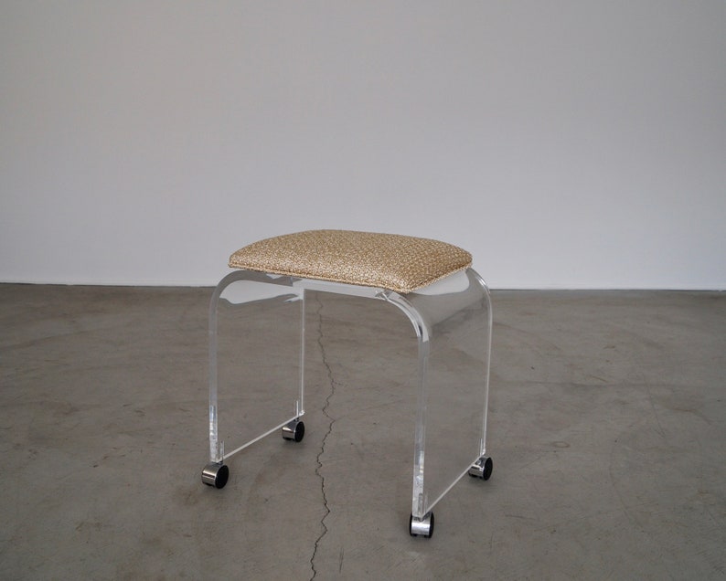 May include: Clear acrylic stool with a rectangular, cushioned seat in a speckled tan fabric. The stool has a curved design and sits on four small wheels for easy movement. A modern piece of furniture.
