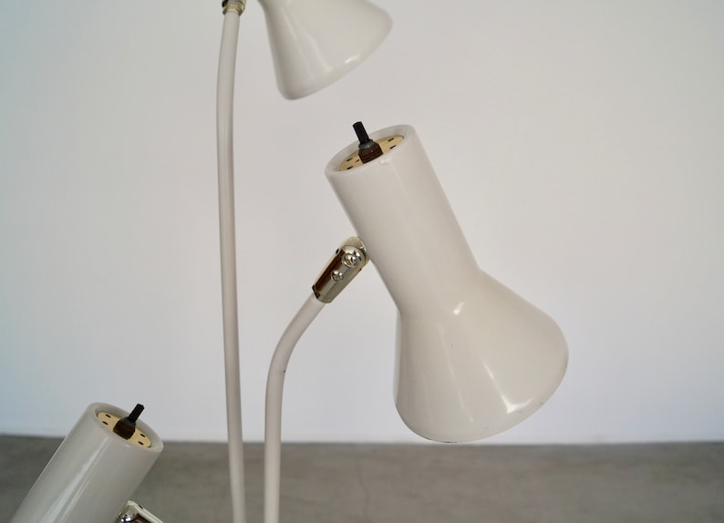 May include: A white, multi-head desk lamp with three adjustable spotlights. The lamp features a slender, curved stem and cone-shaped shades. The lamp is made of metal and has a minimalist design.