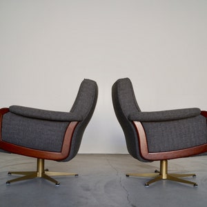 May include: Two gray upholstered armchairs with wooden frames and brass-colored star bases. The chairs have a mid-century modern design, with curved armrests and high backs. The chairs are set against a plain white wall.