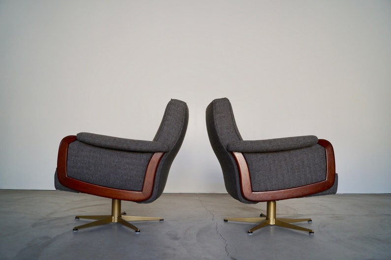 May include: Two gray upholstered armchairs with wooden frames and brass-colored star bases. The chairs have a mid-century modern design, with curved armrests and high backs. The chairs are set against a plain white wall.