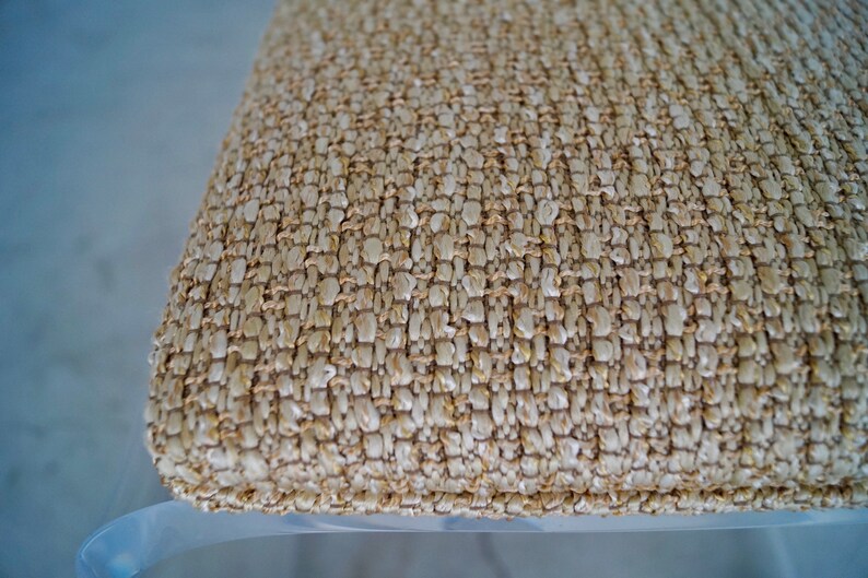 May include: Close-up of a woven cushion with a textured surface. The cushion is made of natural-colored fibers, creating a neutral, earthy tone. The weave pattern is visible, adding to the tactile quality of the design. The cushion appears to be part of a chair or stool.