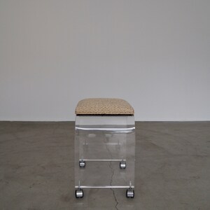 May include: A clear acrylic stool with a rectangular shape and a cushioned seat. The seat has a beige and brown patterned fabric. The stool has four small wheels for mobility. The design is modern and minimalist.